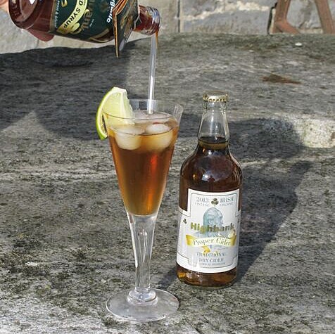 long stemmed glass with straw containing golden liquid,ice and sliced apple beside a bottle of proper cider