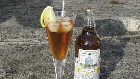 long stemmed glass with straw containing golden liquid,ice and sliced apple beside a bottle of proper cider