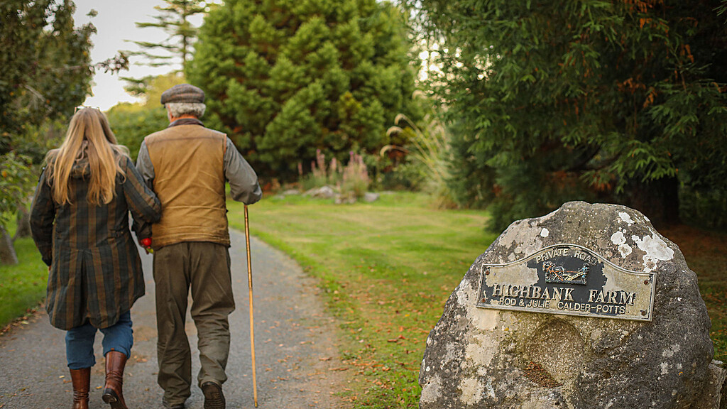 couple walking with a walking stick down Highbank avenue with their backs to you. On the right is a round stone with the Highbank Orchards sign on it.