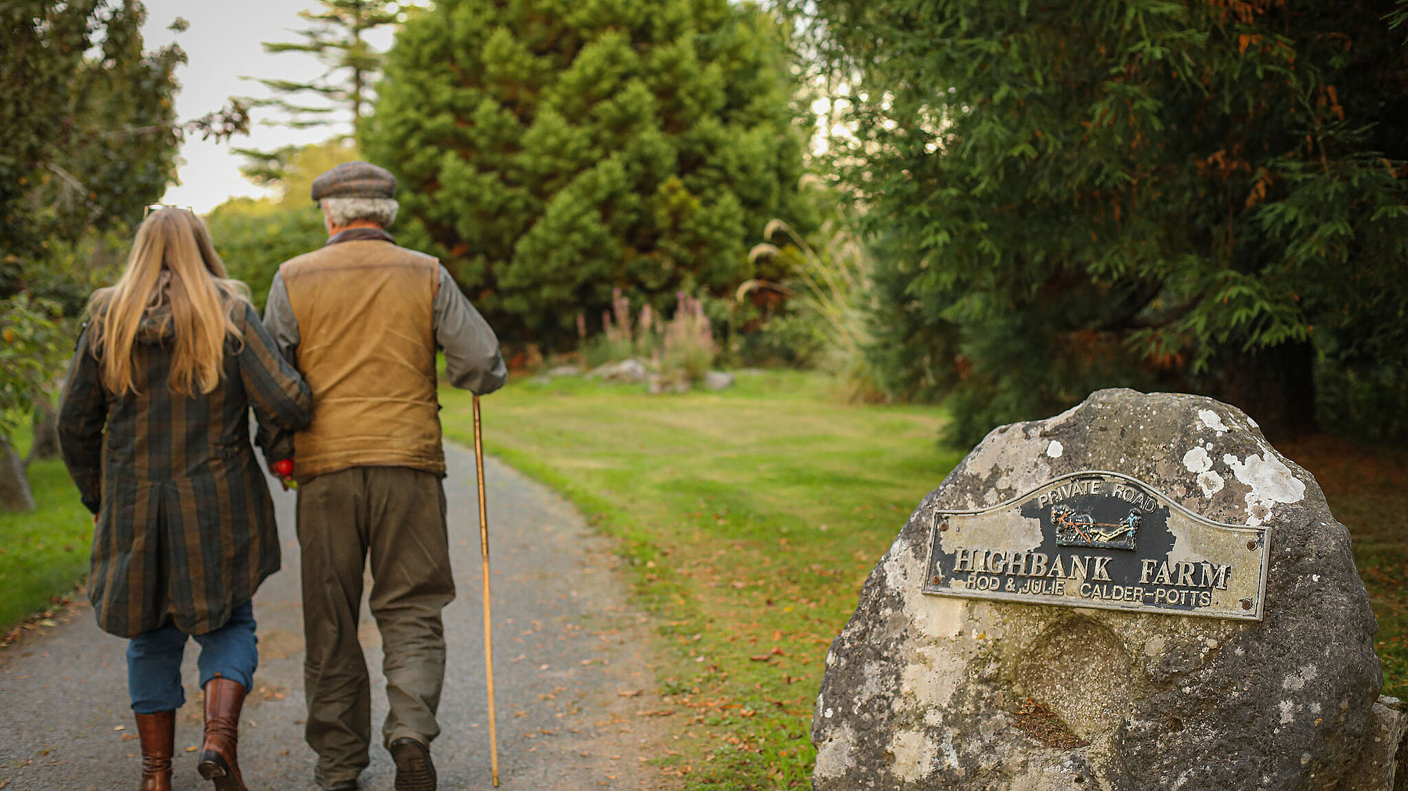 couple walking with a walking stick down Highbank avenue with their backs to you. On the right is a round stone with the Highbank Orchards sign on it.