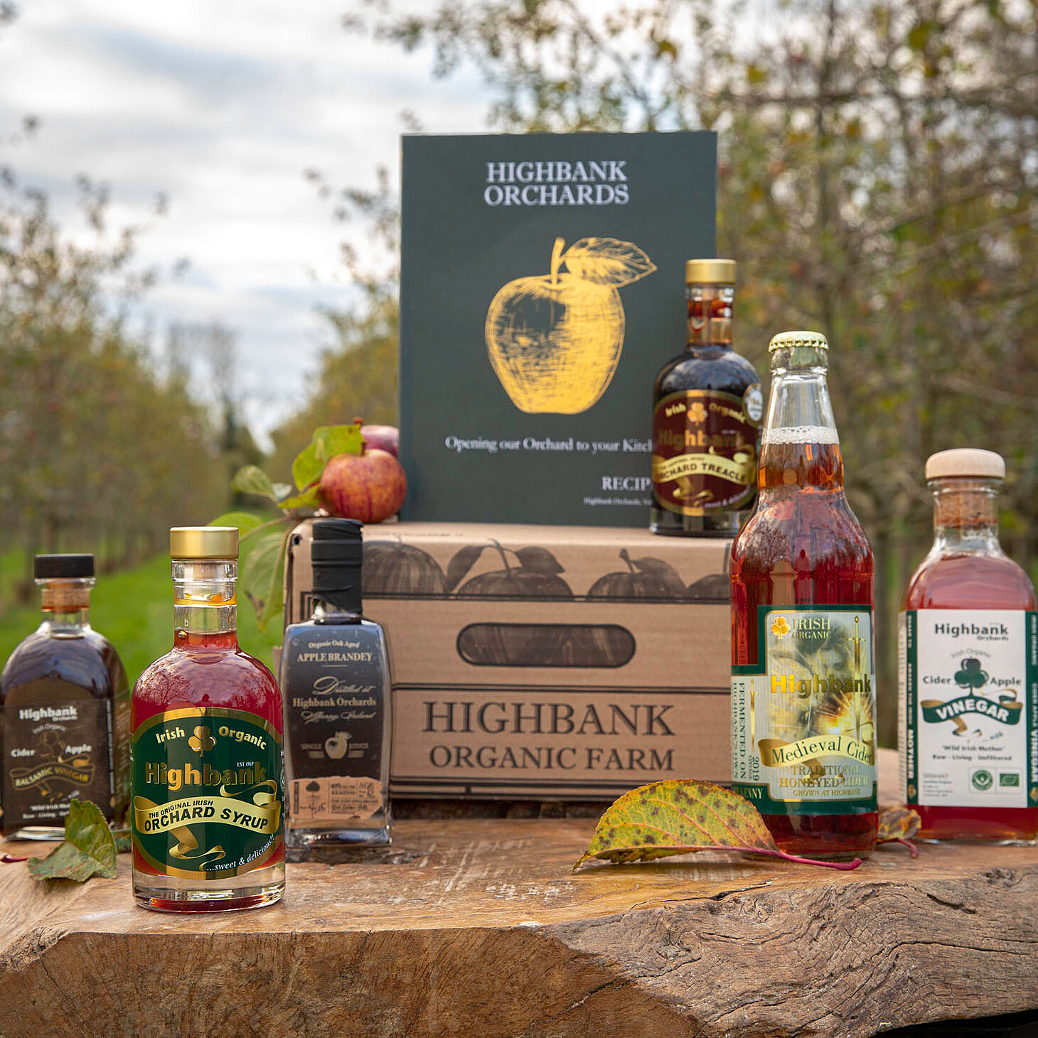 hamper box with hamper contents displayed beside it all on a piece of wood in the orchards. The Highbank recipe book is central on the box with a large golden apple on the cover
