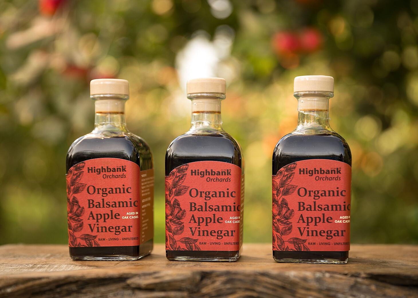 3 bottles of 250ml oak aged apple balsamic vinegar with red labels standing on wood in the orchard