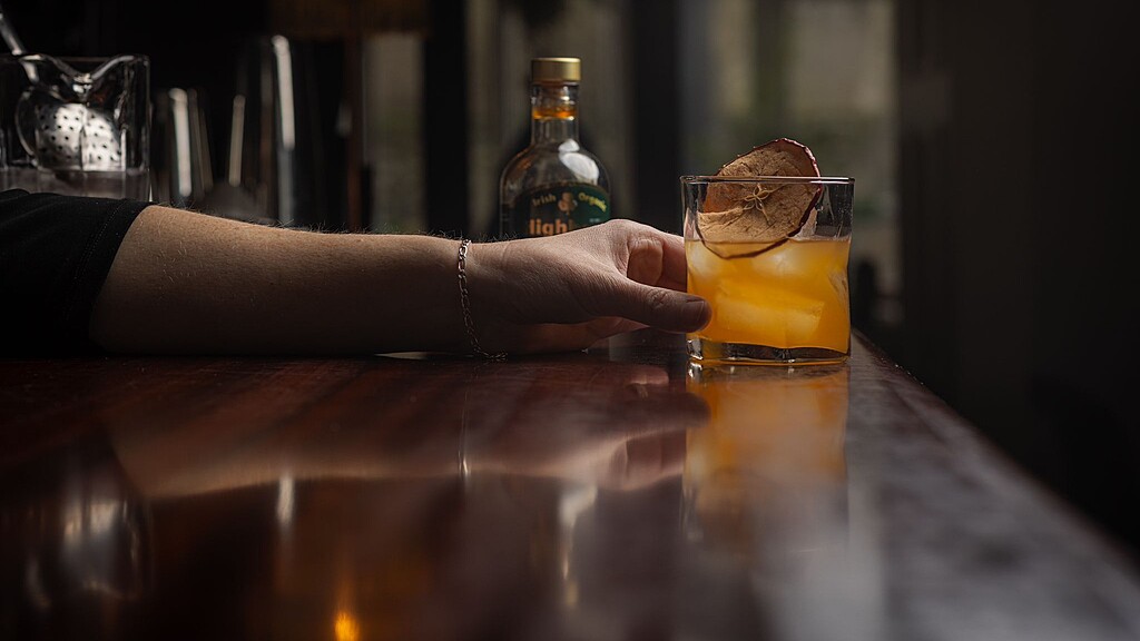 hand on counter holding glass with slice of dried apple and full of orange cocktail