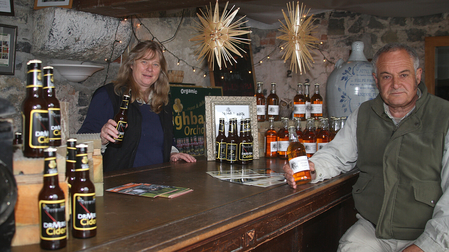 inside farm shop. girl behind wooden counter man on stool drinking Drivers Cider