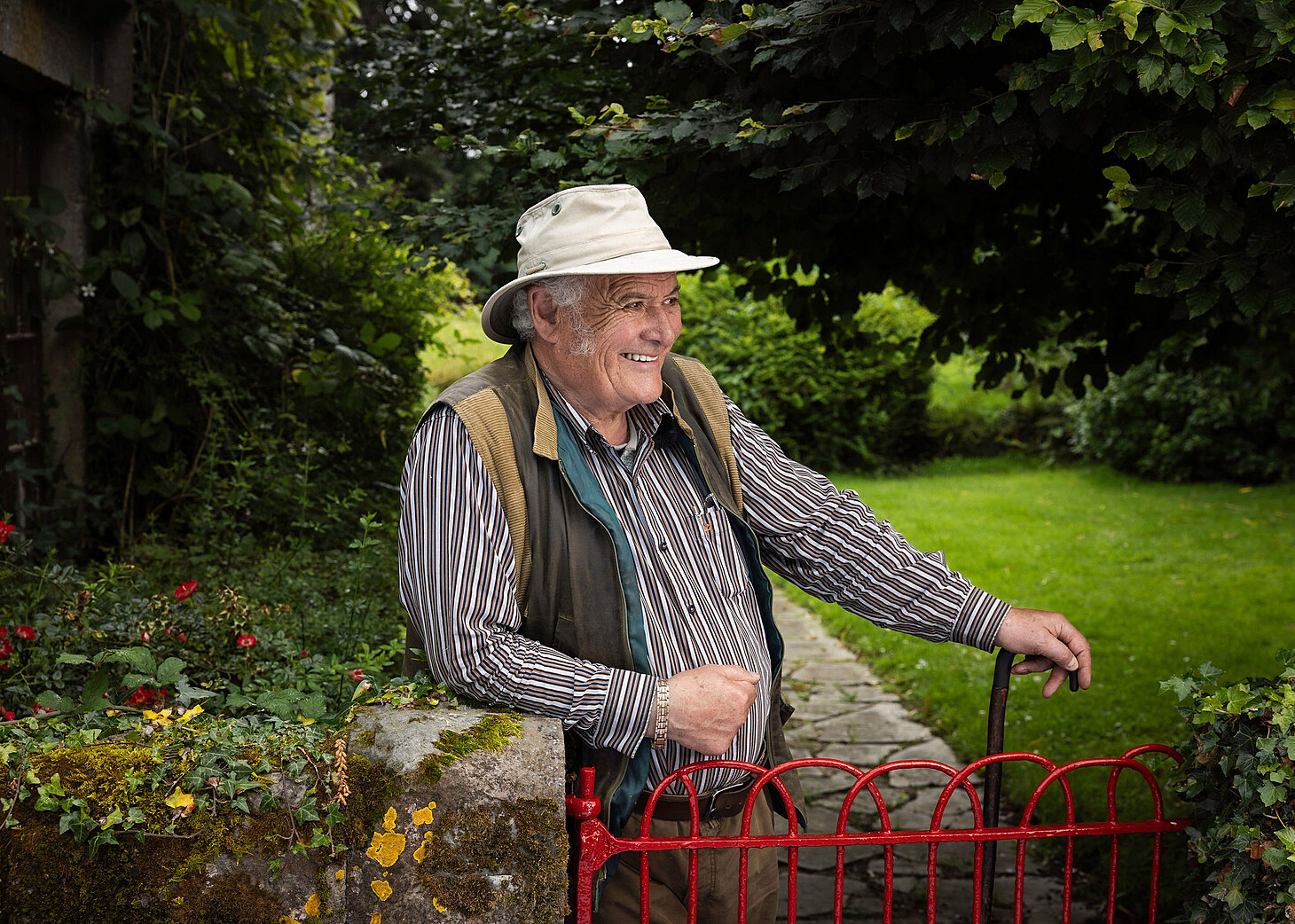 farmer smiling with hat on looking over red gate at front of the house