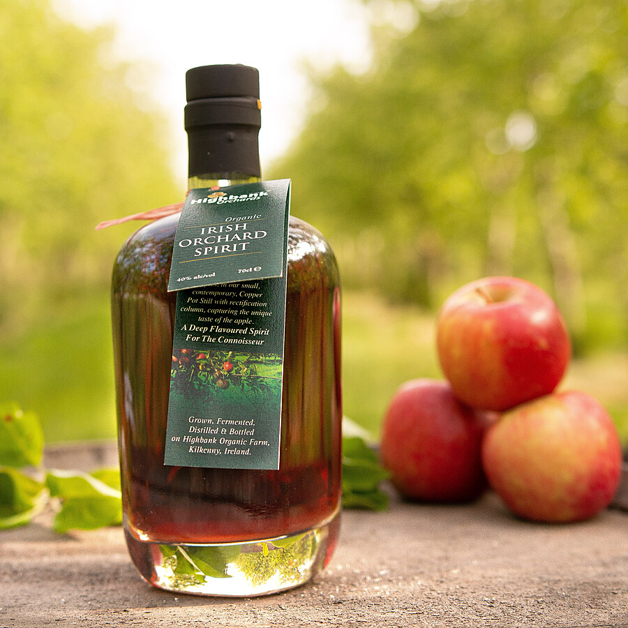 A 700ml round glass bottle with a black top full of Highbank golden Orchard Spirit on wood in the orchards with three red apples piled up beside it The label is dark green around the neck