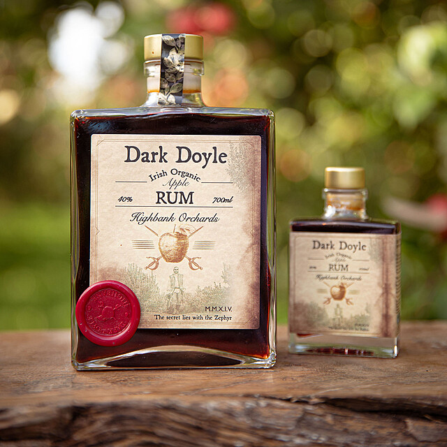 2 x square glass bottles on wood in the orchards. Dark Doyle organic Apple Rum 700ml and small Dark Doyle organic Apple Rum100ml. Label has picture of man carrying bottle out of orchards in pirate outfit. Above him an apple with two crossed swords