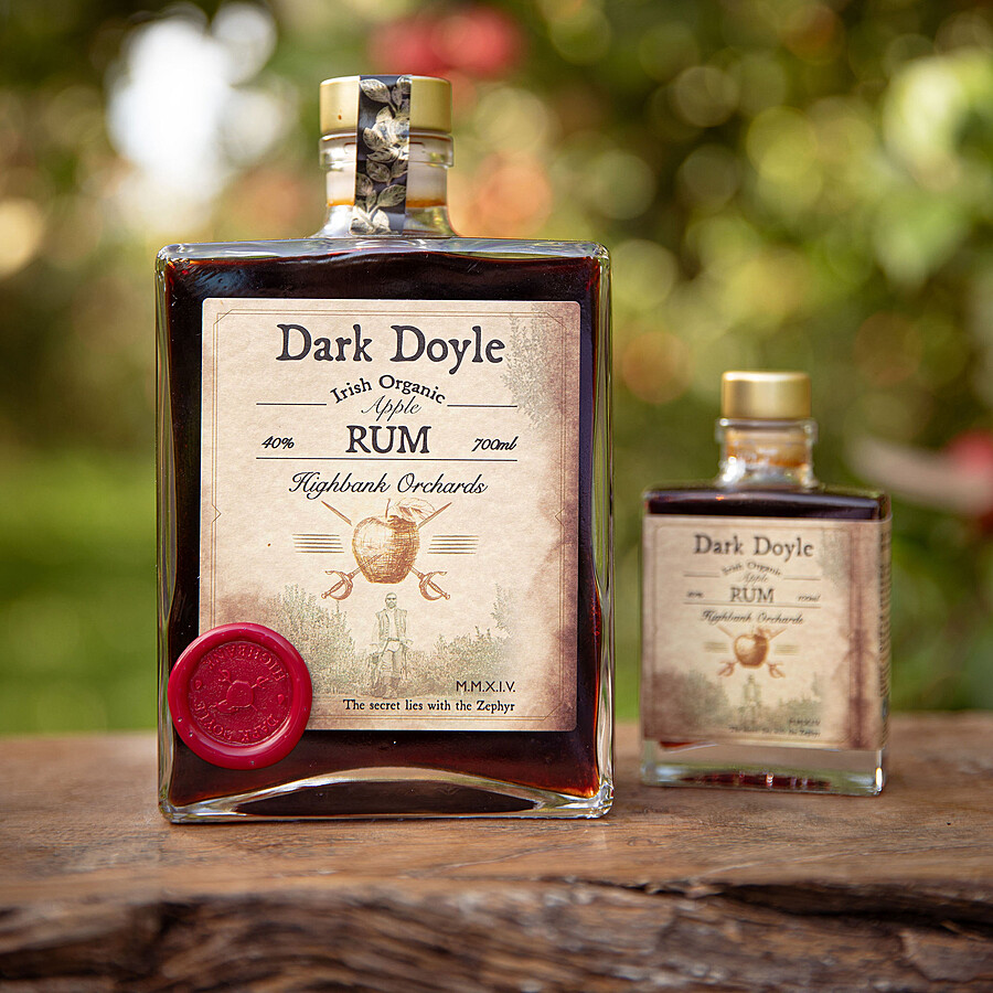 2 x square glass bottles on wood in the orchards. Dark Doyle organic Apple Rum 700ml and small Dark Doyle organic Apple Rum100ml. Label has picture of man carrying bottle out of orchards in pirate outfit. Above him an apple with two crossed swords
