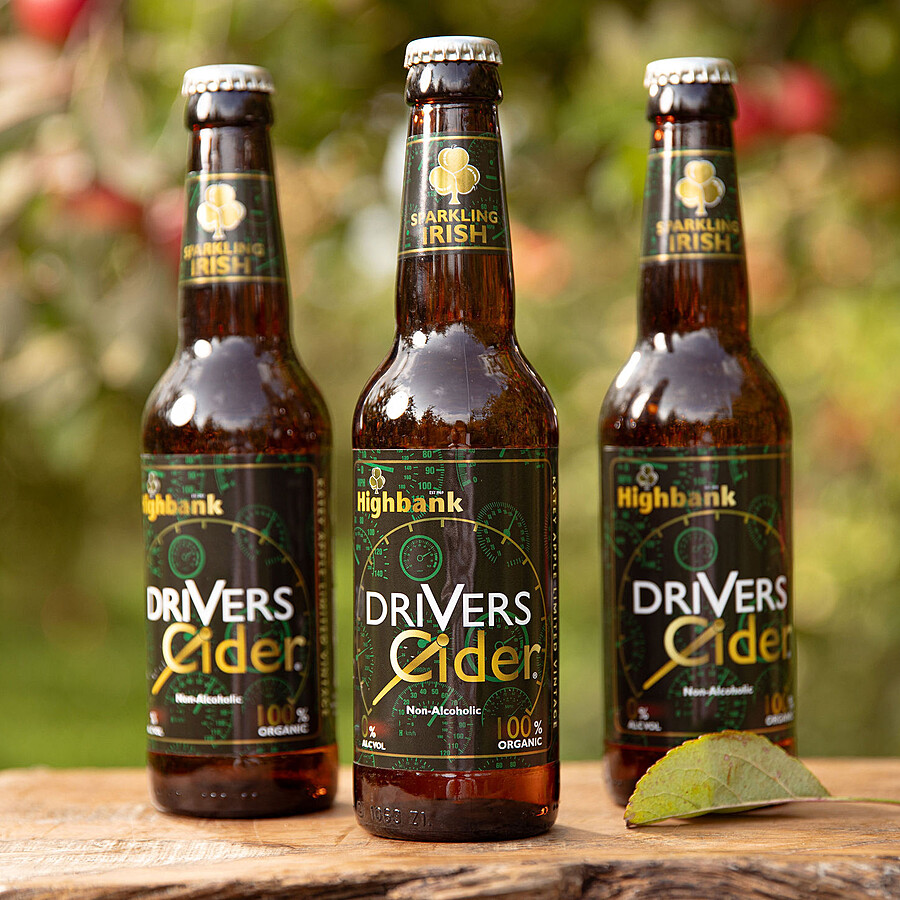 Three black 330ml bottles of Drivers Cider nonalcoholic on wood in the orchards. Labels black with white lettering