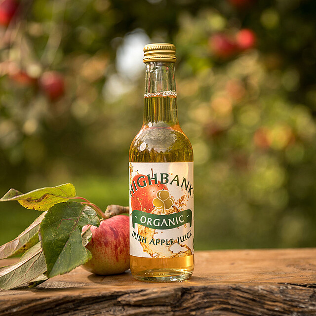 250ml single round gold topped glass bottles full of Highbank golden apple juice on wood in the orchards. White label with large red apple falling from the top left side