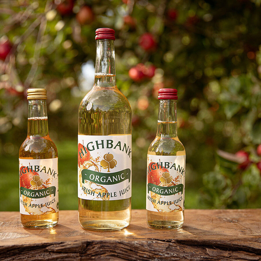 3 round glass bottles full of Highbank golden apple juice on wood in the orchards One large 750ml bottle and two small 250ml bottles either side. The label is white with large red apple falling from the top left side of the label.