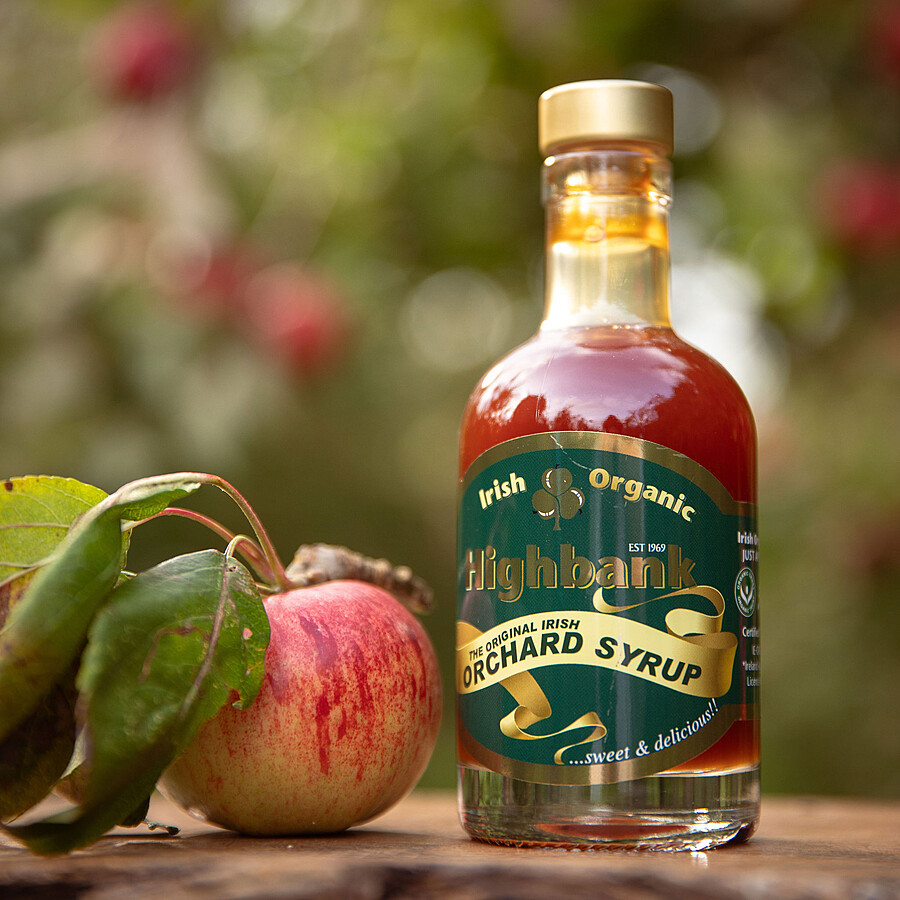 200ml round glass bottle with a gold top of Orchard Syrup standing on wood in the orchards with a red apple with leaves beside it.. The label is dark green with a yellow sash