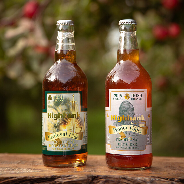 Two 330ml glass bottles of cider in the orchards, One label reads Medieval cider with a girl and horse, the other label reads Proper Cider with a drawing of Henry Flood