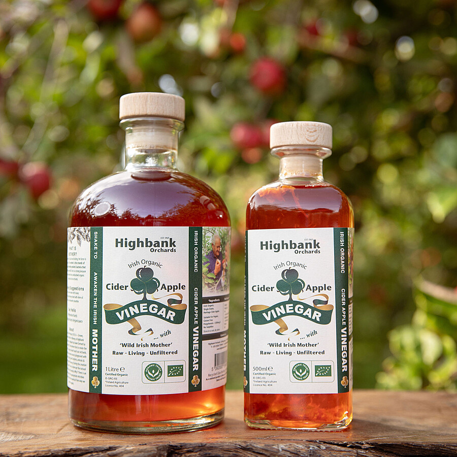 2 x glass bottles on wood in the orchard of Organic Apple Cider Vinegar with Wild Mother. One 1 litre large round wooden corked bottle,the other a 500ml square bottle of apple cider vinegar. Both full of deep orange coloured apple cider vinegar with a white label and green lettering