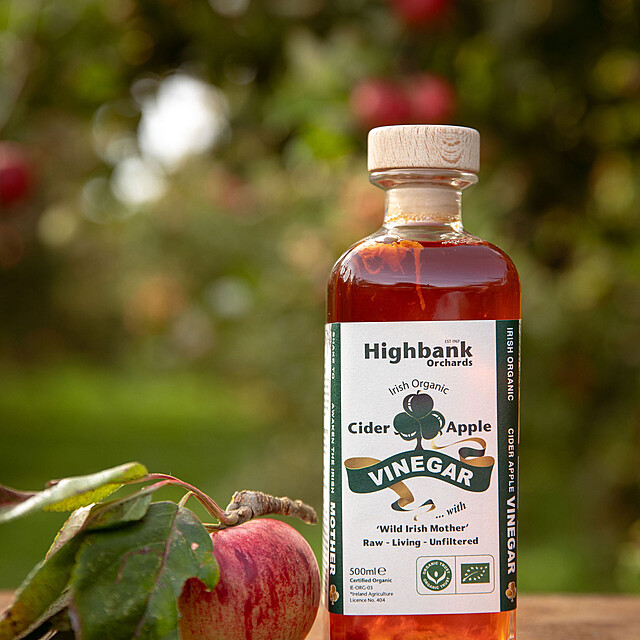500ml square glass bottle of Organic Apple Cider Vinegar with Wild Mother. Full of deep orange coloured with a white label and green lettering. on wood beside an apple and leaves