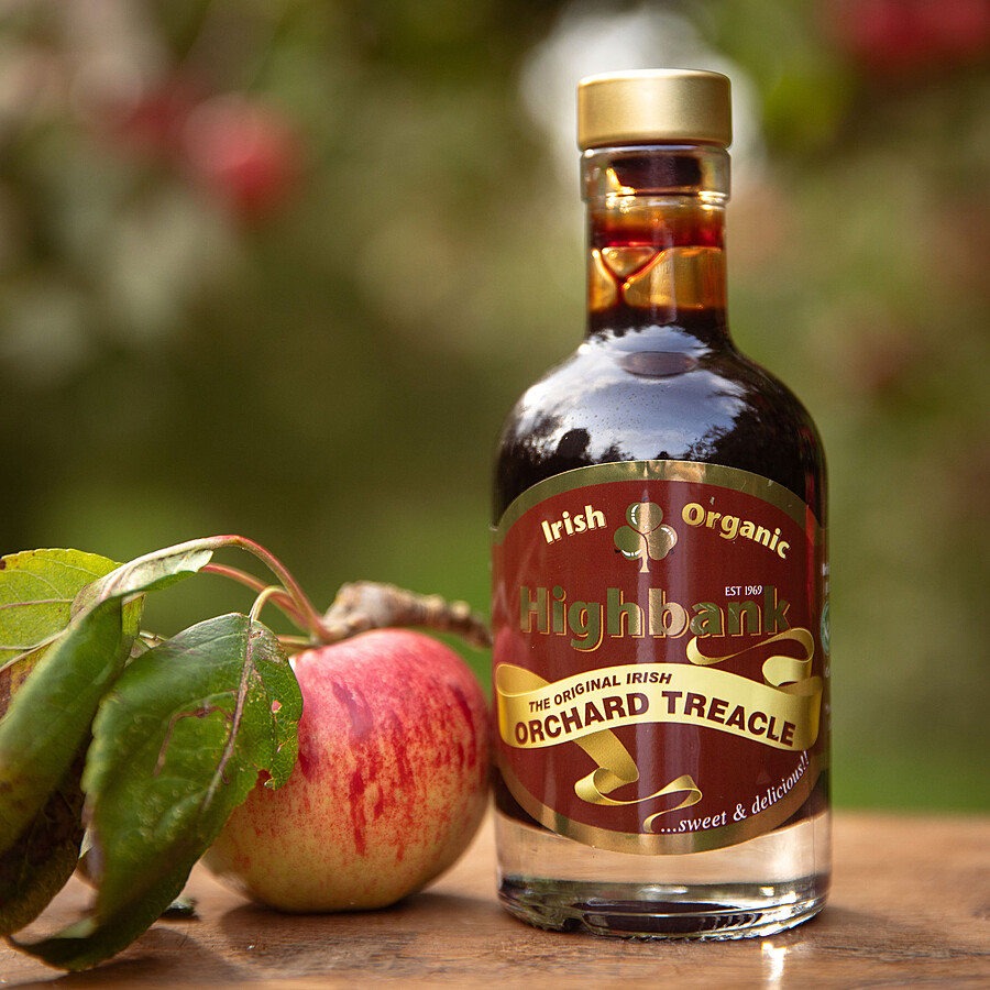 200ml round glass gold topped bottle of Orchard Treacle on wood in the orchards with a red apple and leaves beside it.. Dark Red label with a yellow sash