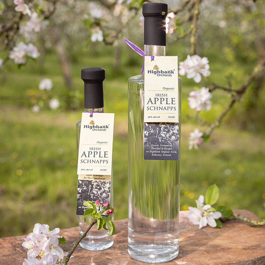 2 x tall glass bottles of Highbank apple Schnapps amid apple blossom 700ml and 200ml bottles side by side on wood