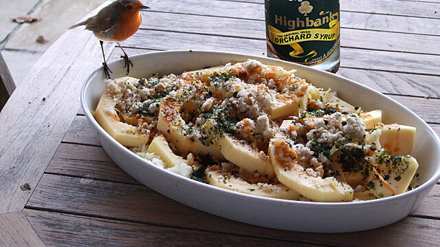 white baking dish with courgettes and breadcrumbs inside and a robin sitting on the edge. a bottle of orchard syrup stands beside the dish on a wooden table