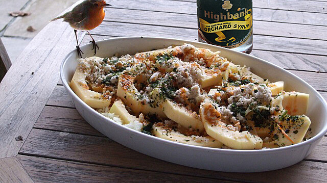 white baking dish with courgettes and breadcrumbs inside and a robin sitting on the edge. a bottle of orchard syrup stands beside the dish on a wooden table